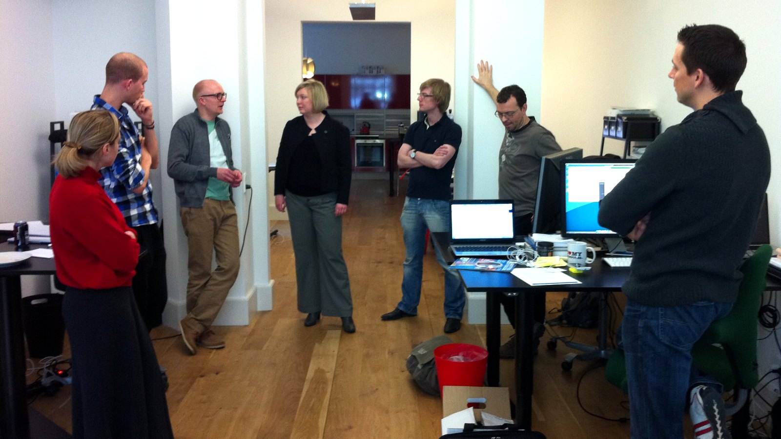 Eight colleagues standing around laptops in an office, holding a daily standup meeting